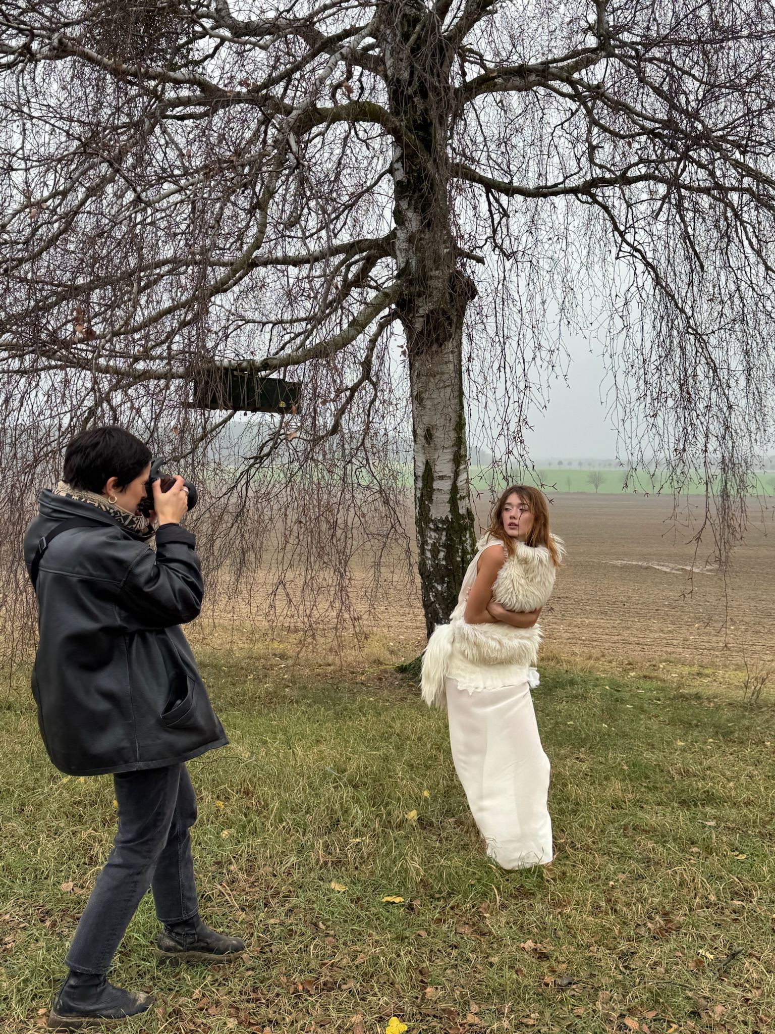 Ein Fotograf richtet die Kamera auf ein Model in weißem Kleid unter einem kahlen Baum.