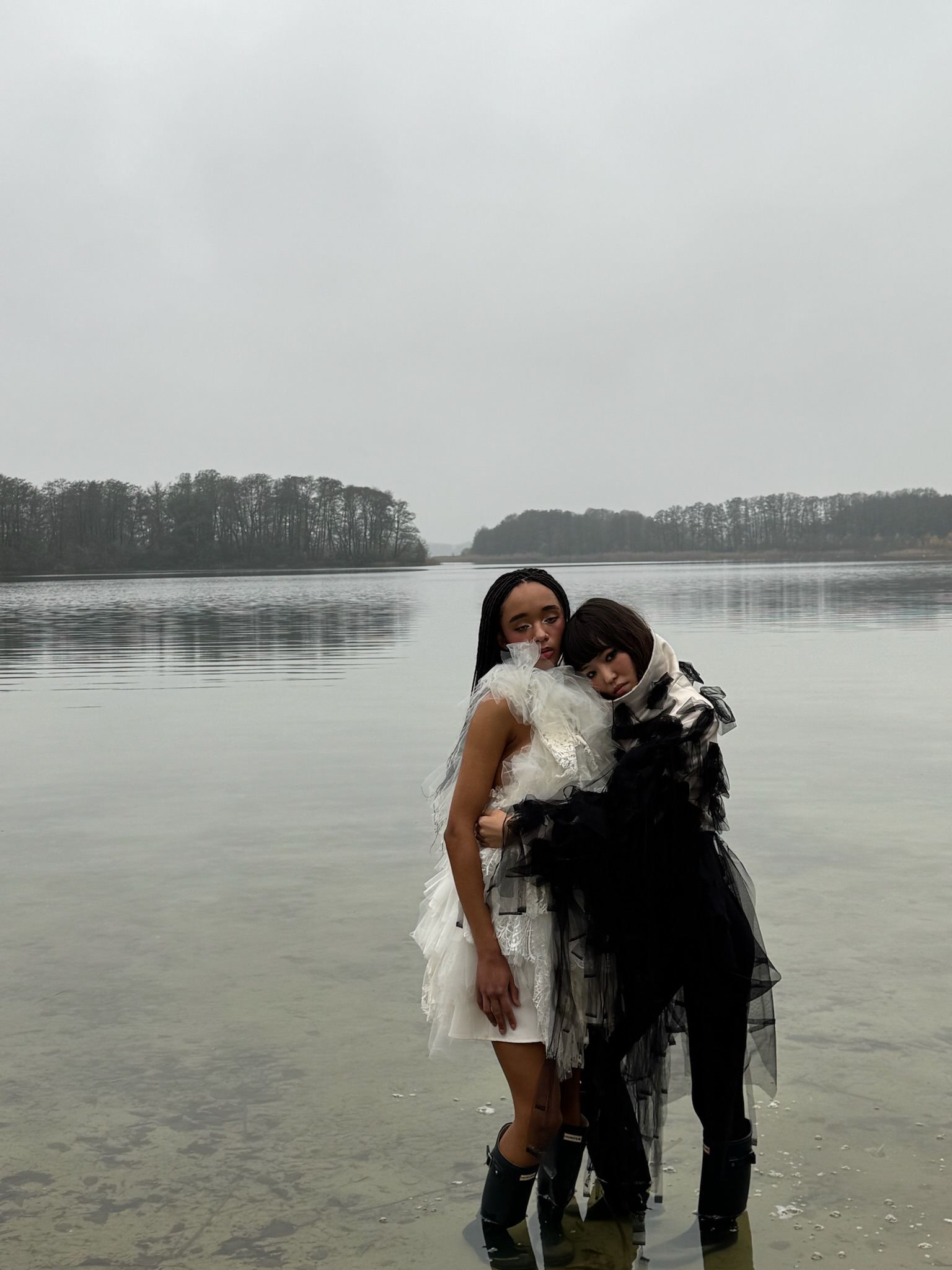 Zwei Models stehen eng beieinander im flachen Wasser eines Sees bei Nebel.