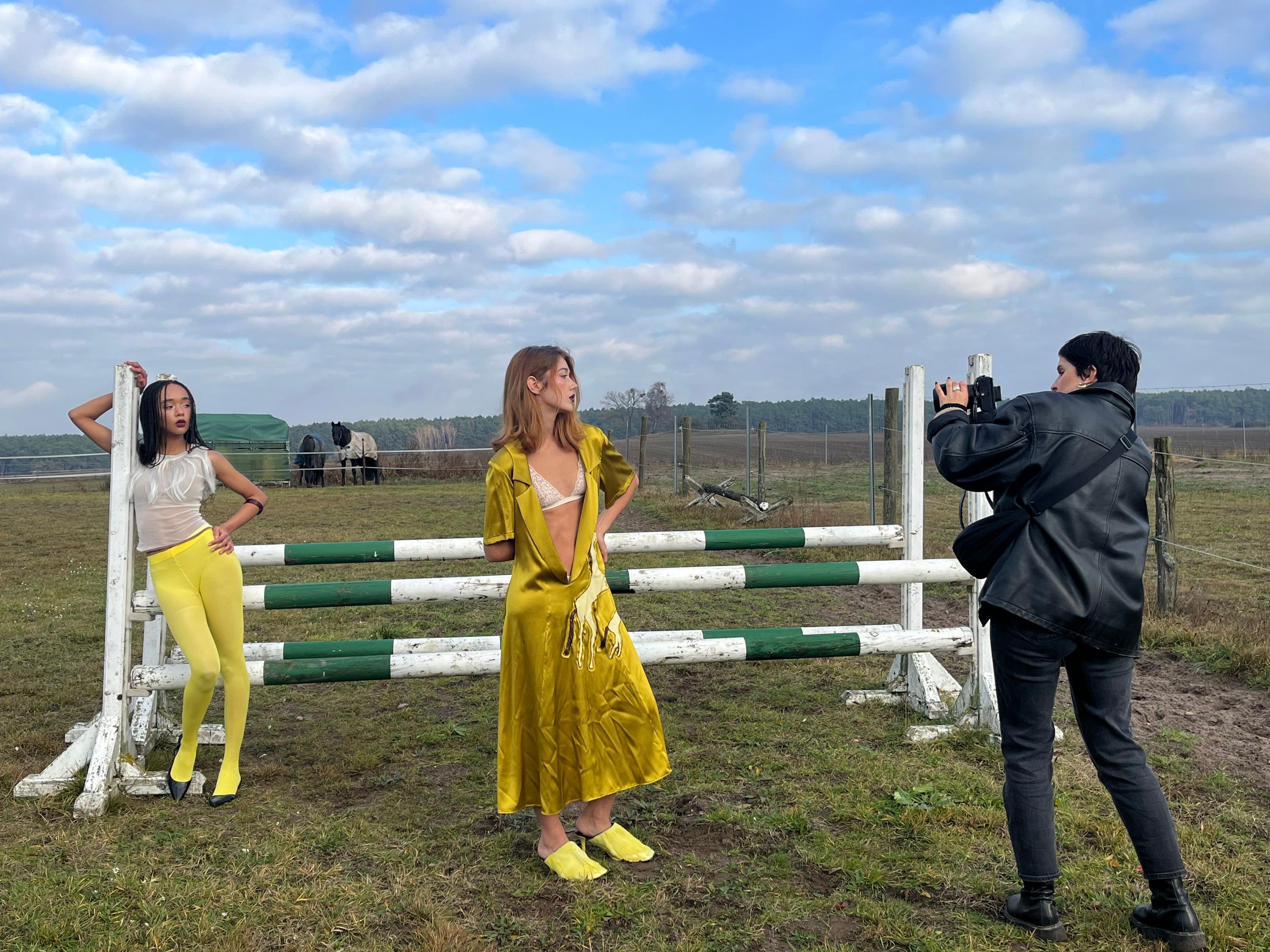 Zwei Models in gelben Looks an einem weißen Sprunghindernis auf der Koppel, dahinter der Fotograf.