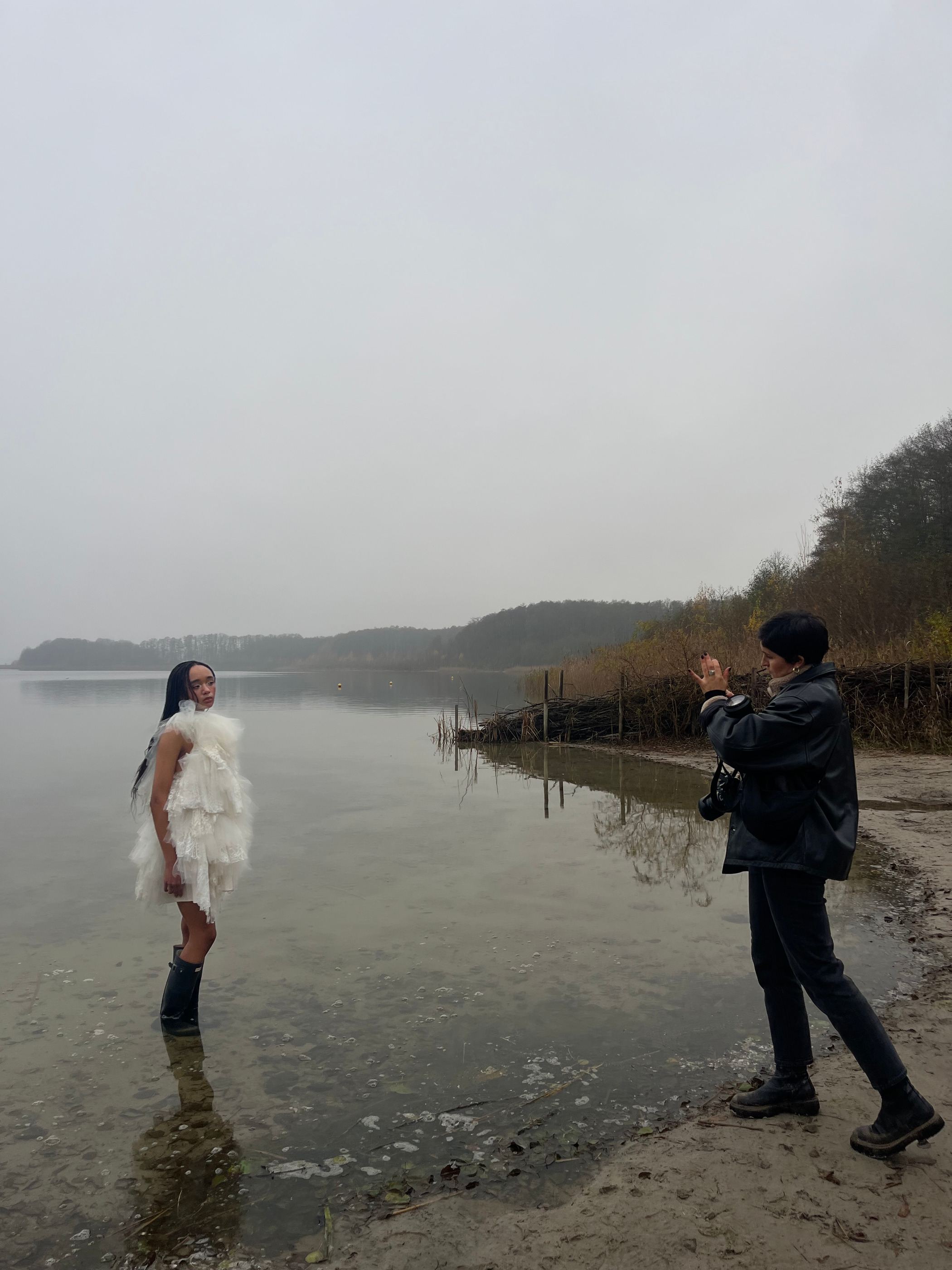Model in hellem Kleid steht im flachen Wasser eines Sees, daneben eine Person in Schwarz.