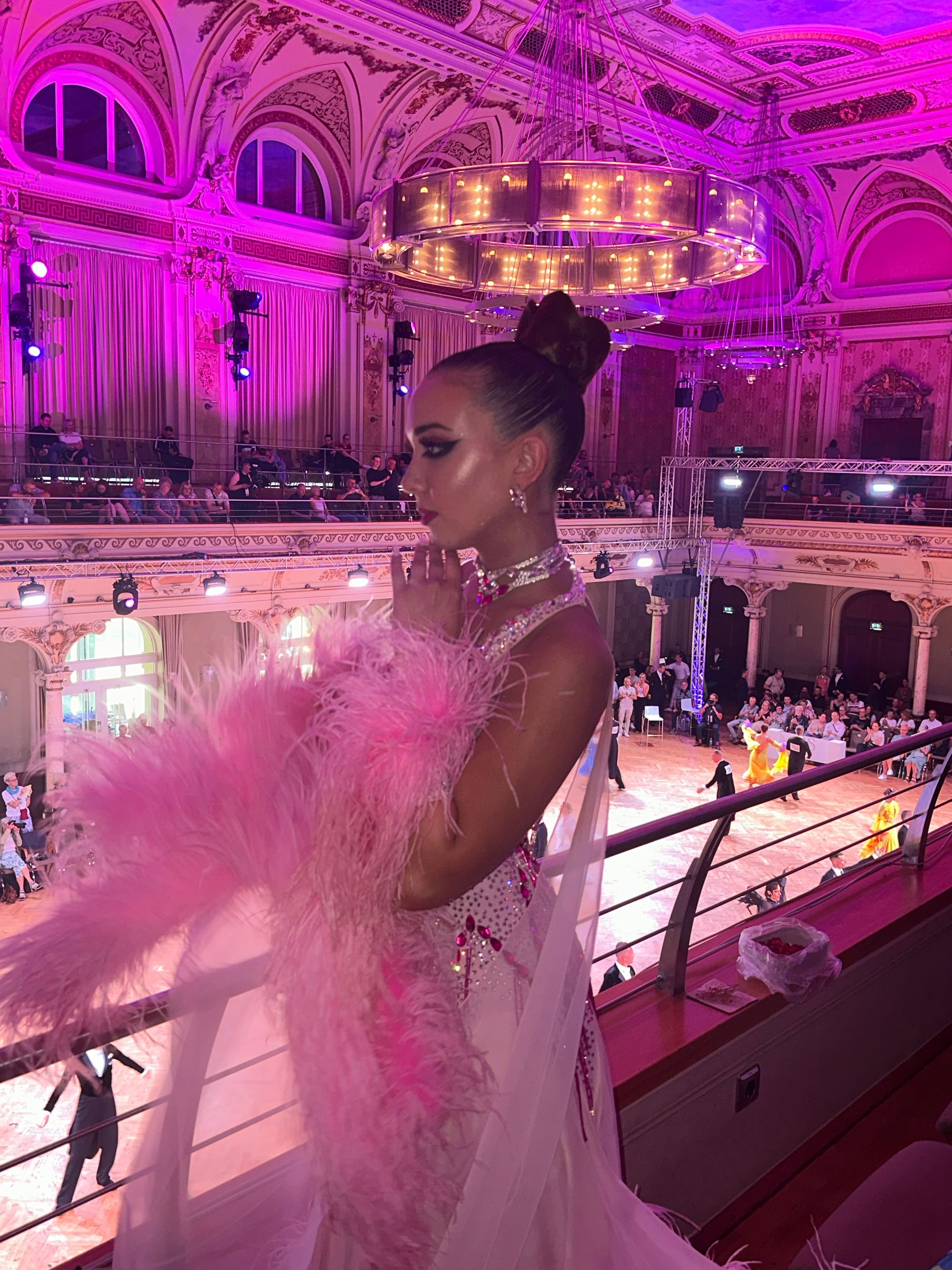 Turnierfläche in einem historischen Ballsaal unter magentafarbenem Licht, ein Standardkleid mit rosa Federn schwingt aus.