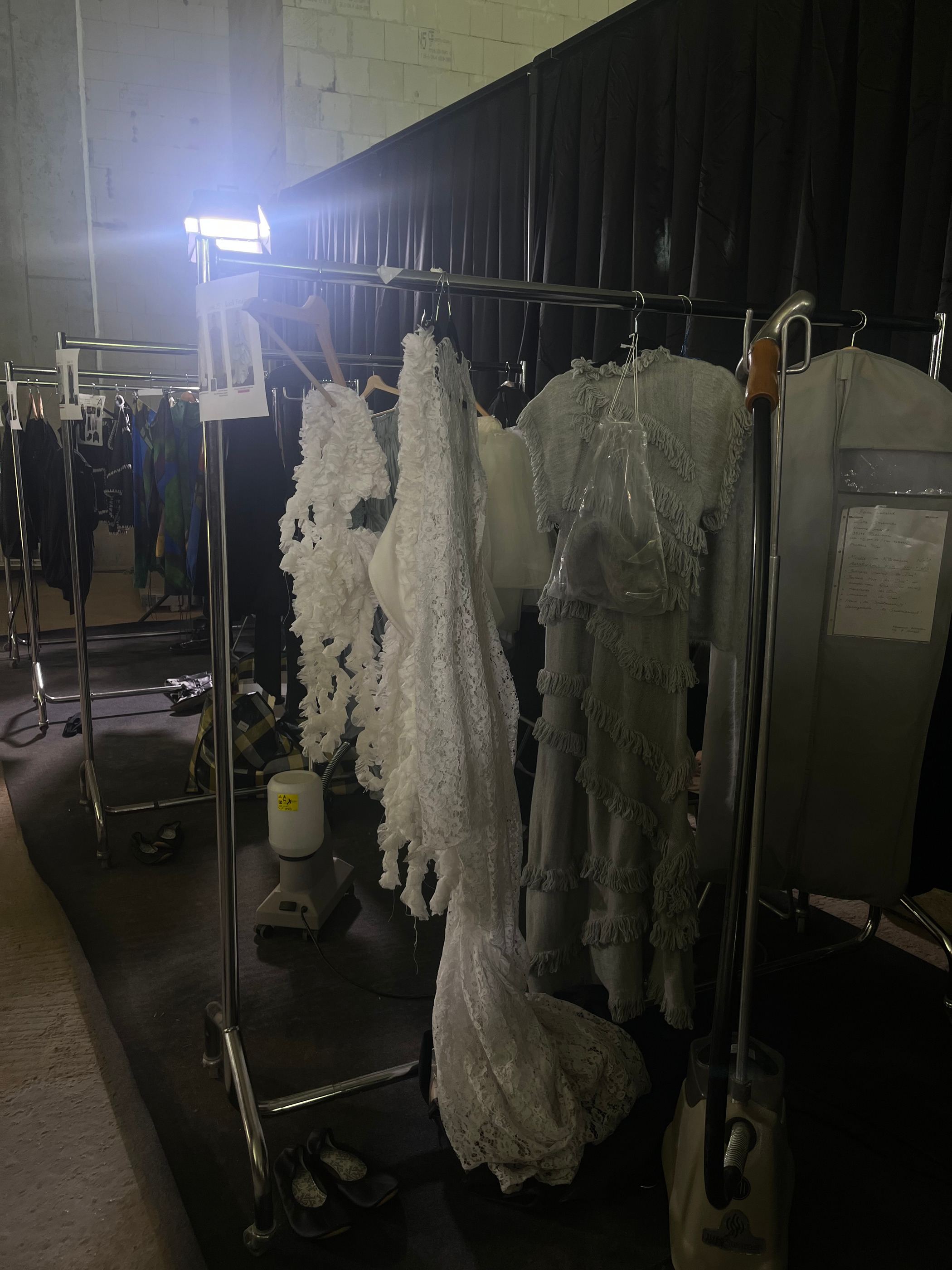 Kleiderstange mit hellen, transparenten Looks im Backstagebereich, angestrahlt von einem Scheinwerfer.