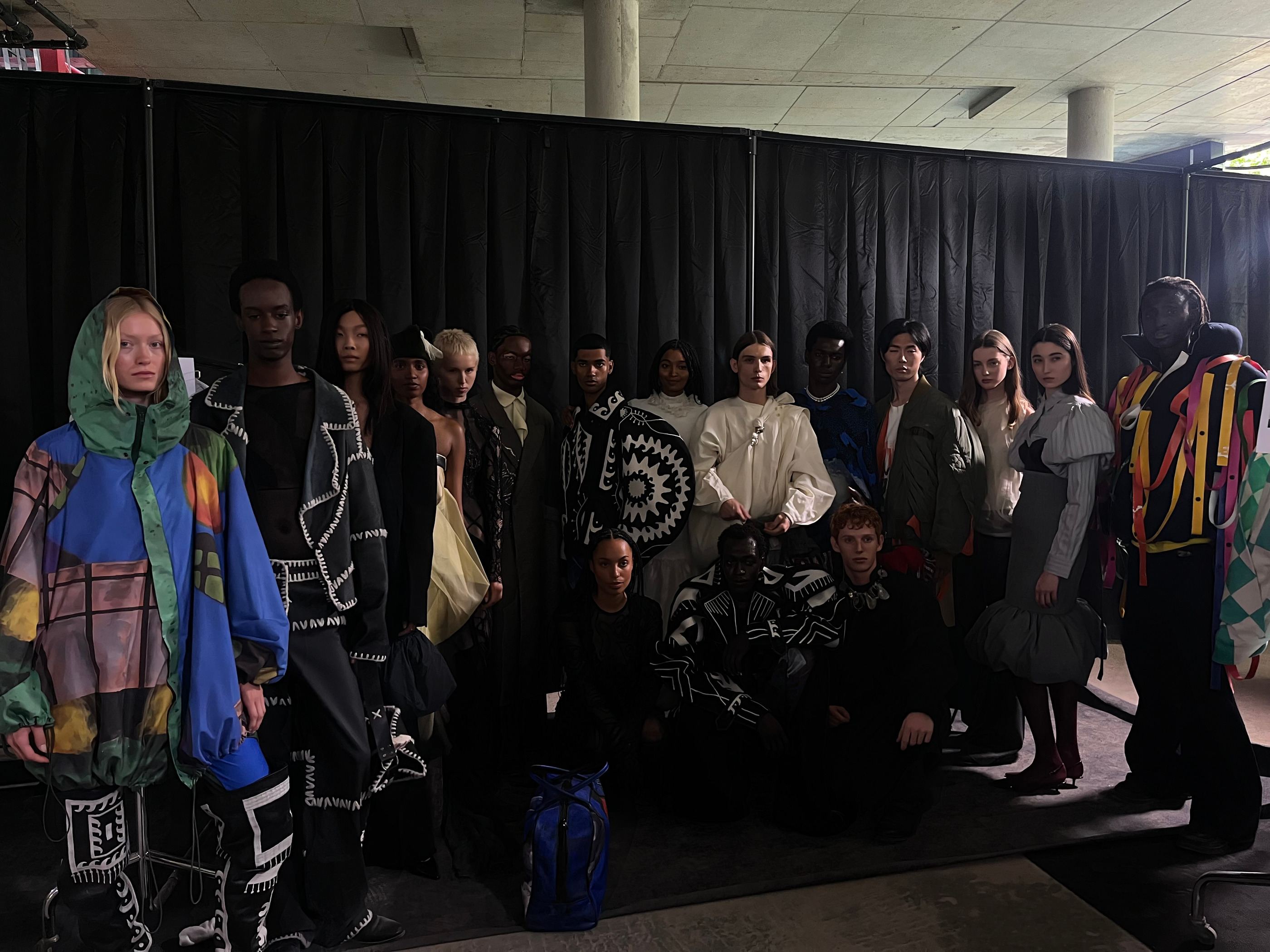 Models in ihren Looks stehen backstage in einer Reihe vor einem schwarzen Vorhang.