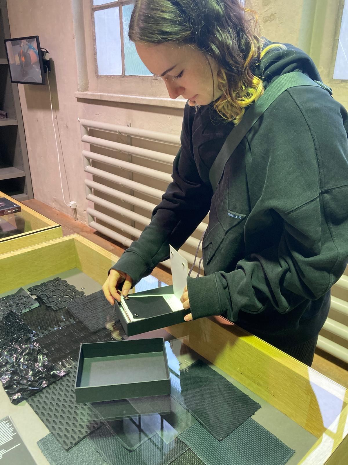 Eine Person legt eine schwarze Materialprobe in einen flachen Kasten der Ausstellung.